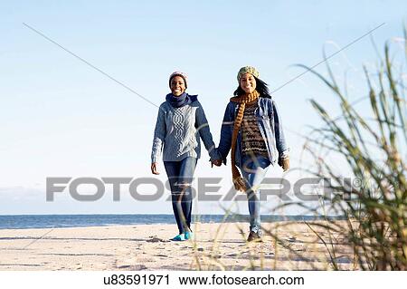 "Two young women, walking along beach, holding hands, smiling" View Large Photo Image Stock Image - "Two young women, walking along beach, holding hands, smiling". Fotosearch