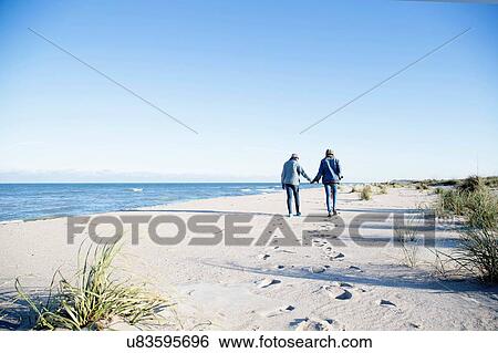 Stock Photograph - "Two young women, walking along beach, holding hands, rear view". Fotosearch
