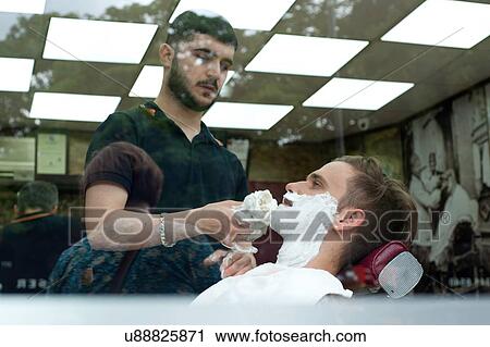 Stock Image - View through window of young man in barbershop applying shaving cream to customers face. Fotosearch