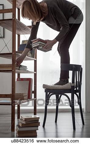 Stock Photograph - Woman standing on chair to arrange books on living room bookshelf. Fotosearch