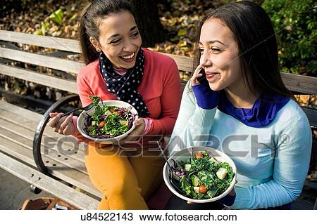 Young adult female twins sitting on park bench chatting on smartphone and eating lunch View Large Photo Image Stock Image - Young adult female twins sitting on park bench chatting on smartphone and eating lunch. Fotosearch