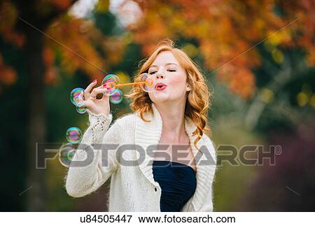 Stock Photo - Young beautiful woman with red wavy hair blowing bubbles in autumn forest. Fotosearch