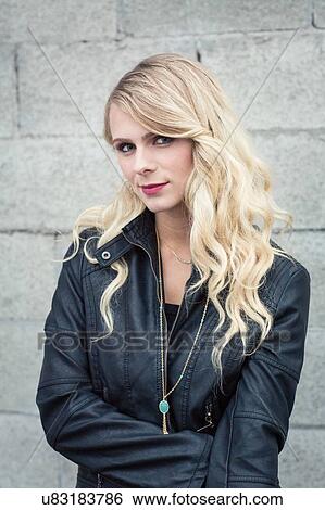 Stock Photograph - Young blonde haired woman in front of breeze block wall wearing leather jacket looking at camera smiling. Fotosearch