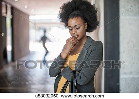 Stock Image - Young businesswomen with hand on chin leaning against office wall. Fotosearch