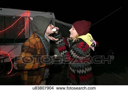 Picture - "Young couple at camp site, at night, young woman feeding young man, laughing". Fotosearch