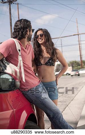 Stock Photography - Young couple chatting whilst leaning against red car at roadside, Los Angeles, California, USA. Fotosearch
