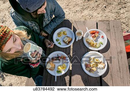 Stock Image - "Young couple eating lunch at picnic table, elevated view". Fotosearch