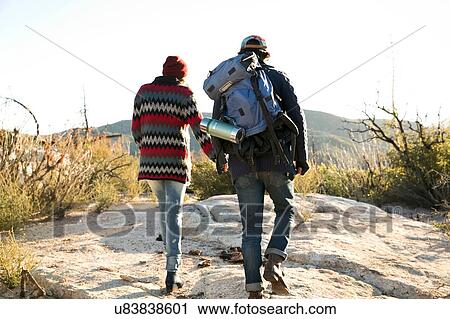 Stock Image - "Young couple hiking in rural setting, holding hands, rear view". Fotosearch