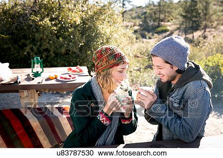 Stock Image - "Young couple in rural setting, sitting at picnic bench, drinking coffee". Fotosearch