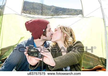 Stock Photography - "Young couple sitting in tent, kissing, taking self portrait using camera". Fotosearch