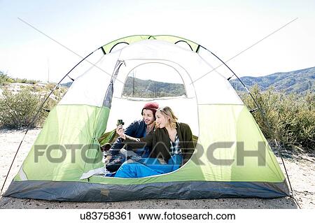 Stock Image - "Young couple sitting in tent, taking self portrait using camera". Fotosearch
