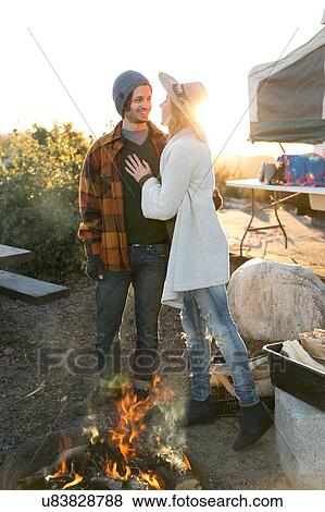 Stock Photo - "Young couple standing by camping fire, face to face, smiling". Fotosearch