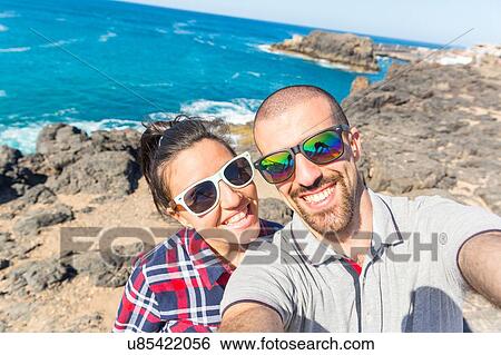 Stock Photograph - Young couple taking smartphone selfie at sea, El Cotillo, Fuerteventura, Spain. Fotosearch