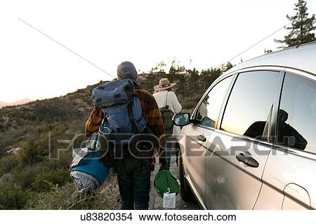 Picture - "Young couple walking away from parked car carrying camping equipment, rear view". Fotosearch