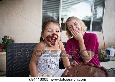 Stock Photography - "Young girl, chocolate cake on her face, sitting with family member, laughing". Fotosearch