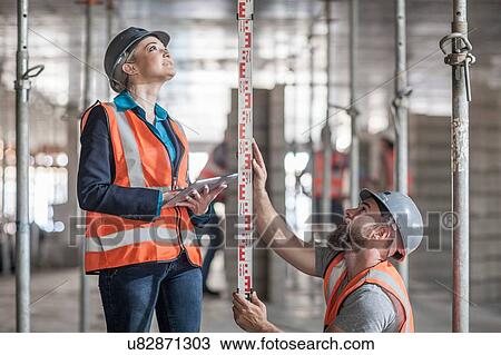 Stock Image - Young male and female builders measuring construction with ruler. Fotosearch
