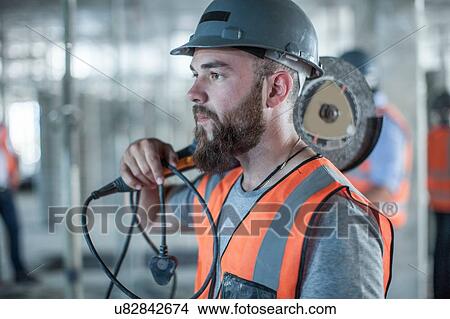 Picture - Young male builder carrying circular saw on shoulder on construction site. Fotosearch