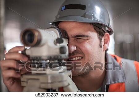 Stock Photo - Young male surveyor looking through theodolite on construction site. Fotosearch