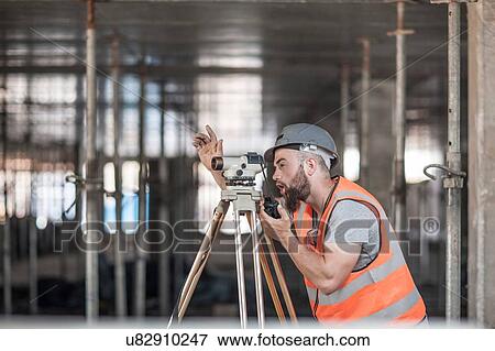 Stock Photo - Young male surveyor looking through theodolite giving hand signal on construction site. Fotosearch