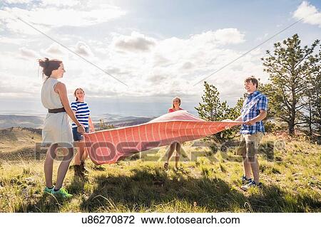 Stock Image - Young man and female friends preparing picnic blanket on hill, Bridger, Montana, USA. Fotosearch