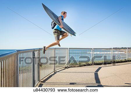 "Young man, carrying surfboard, jumping from railings beside sea" View Large Photo Image Stock Image - "Young man, carrying surfboard, jumping from railings beside sea". Fotosearch