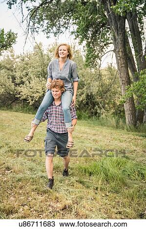 Stock Image - Young man carrying teenage girl on shoulders in rural field. Fotosearch