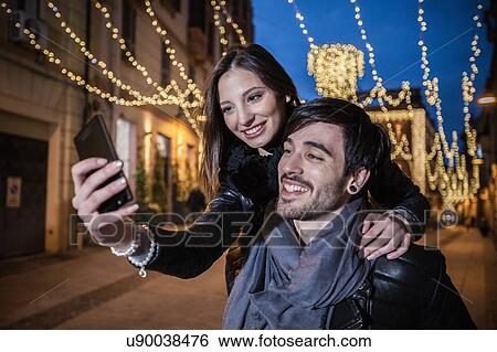 Stock Photograph - Young man giving young woman piggyback using smartphone to take selfie, smiling. Fotosearch