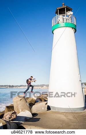 Stock Photo - "Young man holding skateboard, jumping off of breakwater, beside lighthouse". Fotosearch