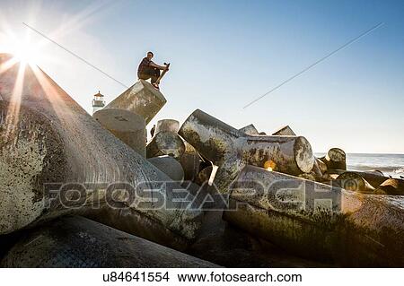 Picture - "Young man holding skateboard, sitting on breakwater, looking towards ocean". Fotosearch