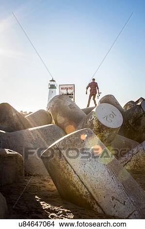 "Young man holding skateboard, standing on breakwater near lighthouse, rear view" View Large Photo Image Picture - "Young man holding skateboard, standing on breakwater near lighthouse, rear view". Fotosearch