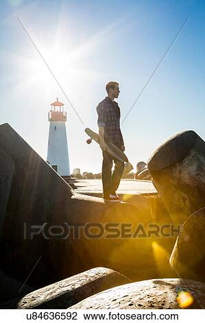 Stock Image - "Young man holding skateboard, standing on breakwater near lighthouse". Fotosearch
