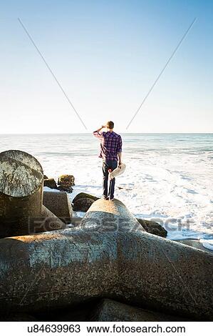 "Young man holding skateboard, standing on breakwater, looking towards ocean, rear view" View Large Photo Image Stock Image - "Young man holding skateboard, standing on breakwater, looking towards ocean, rear view". Fotosearch