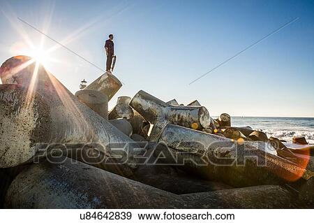 "Young man holding skateboard, standing on breakwater, looking towards ocean" View Large Photo Image Stock Photo - "Young man holding skateboard, standing on breakwater, looking towards ocean". Fotosearch