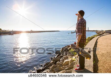 "Young man, holding skateboard, standing at water's edge, looking at view" View Large Photo Image Stock Photograph - "Young man, holding skateboard, standing at water's edge, looking at view". Fotosearch