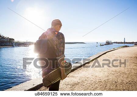 Stock Photography - "Young man, holding skateboard, walking along water's edge, rear view". Fotosearch
