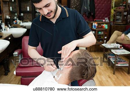 Stock Image - Young man in barbershop looking down shaving customer with straight razor. Fotosearch