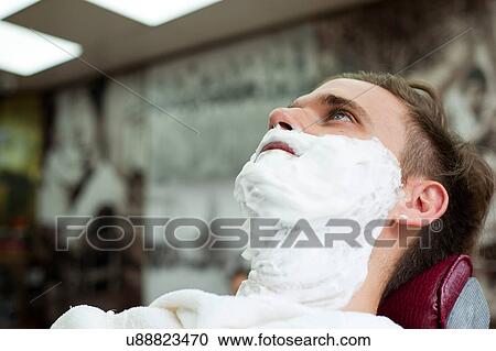 Stock Image - Young man in barbershop with face covered in shaving cream looking up. Fotosearch