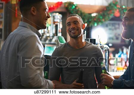 Stock Image - Young man in public house holding beer bottle looking at friend smiling. Fotosearch