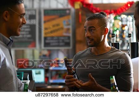 Stock Photo - Young man in public house holding beer bottle talking to friend. Fotosearch
