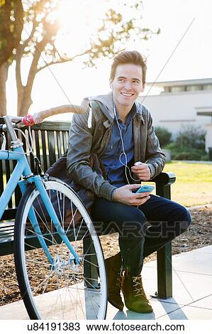 Young man listening to earphone music on sunlit park bench View Large Photo Image Stock Image - Young man listening to earphone music on sunlit park bench. Fotosearch