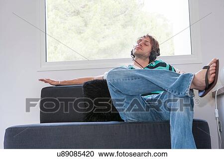 Stock Image - Young man on living room sofa listening to music on headphones. Fotosearch