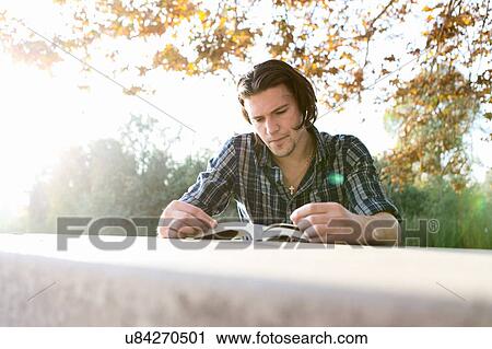 Stock Image - Young man outdoors sitting at table looking down reading book. Fotosearch