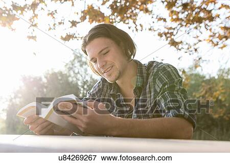 Stock Photo - "Young man outdoors sitting at table looking down reading book, smiling". Fotosearch