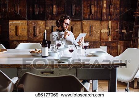 Young man reading digital tablet at dining table View Large Photo Image Stock Photo - Young man reading digital tablet at dining table. Fotosearch