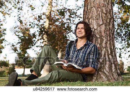 Stock Photo - "Young man sitting against tree holding book, looking away smiling". Fotosearch
