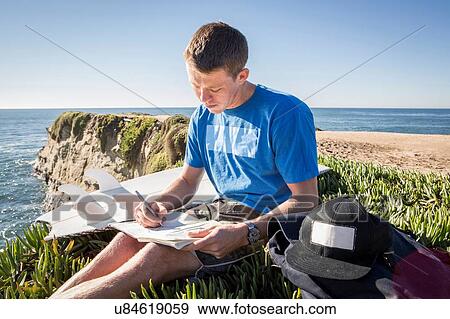Stock Photo - "Young man sitting by cliff edge, writing in book". Fotosearch