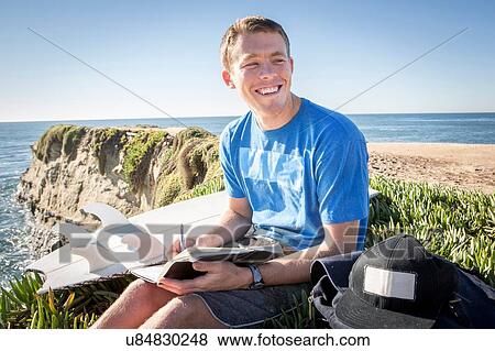 "Young man sitting by cliff edge, writing in book, smiling" View Large Photo Image Stock Photo - "Young man sitting by cliff edge, writing in book, smiling". Fotosearch