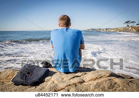 Stock Image - "Young man sitting by rock, looking out to sea, rear view". Fotosearch