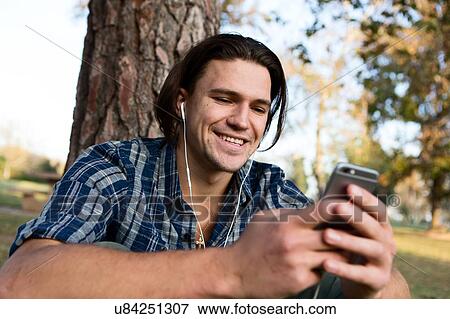 Young man sitting by tree wearing earphones looking at smartphone smiling View Large Photo Image Stock Photo - Young man sitting by tree wearing earphones looking at smartphone smiling. Fotosearch