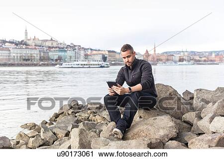 Stock Image - Young man sitting on rocks using digital tablet, Budapest, Hungary. Fotosearch